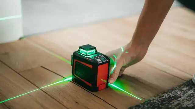 Poziomica laserowa z zielonymi liniami na drewnianej podłodze, obsługiwana przez dłoń.
