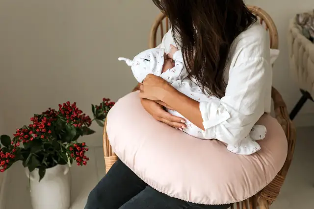 Kobieta siedzi na krześle, trzymając na rękach noworodka. Pokazuje, jak karmić noworodka, otulając go delikatnie.