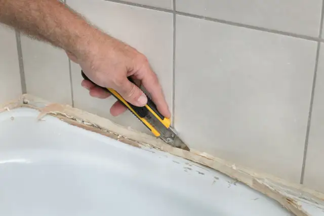 Dłoń z nożem odcina stary silikon z wanny.
