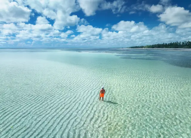 Mężczyzna w pomarańczowych spodenkach stoi w płytkiej, turkusowej wodzie na tle piaszczystego dna i błękitnego nieba z chmurami.