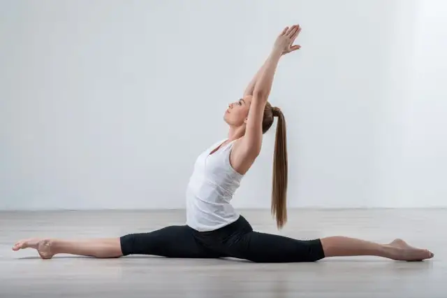 Kobieta w białej koszulce i czarnych legginsach wykonuje szpagat z rękami uniesionymi nad głową.