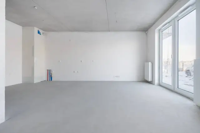 Pusta przestrzeń w stanie deweloperskim: betonowa posadzka, białe ściany, instalacje wodne i elektryczne, grzejnik, duże okna.