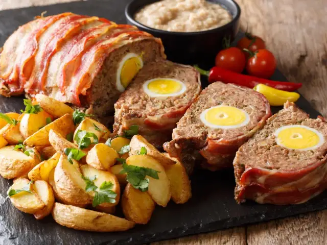 Pyszny klops z jajkiem w środku, zawinięty w boczek, podany z pieczonymi ziemniakami i warzywami.