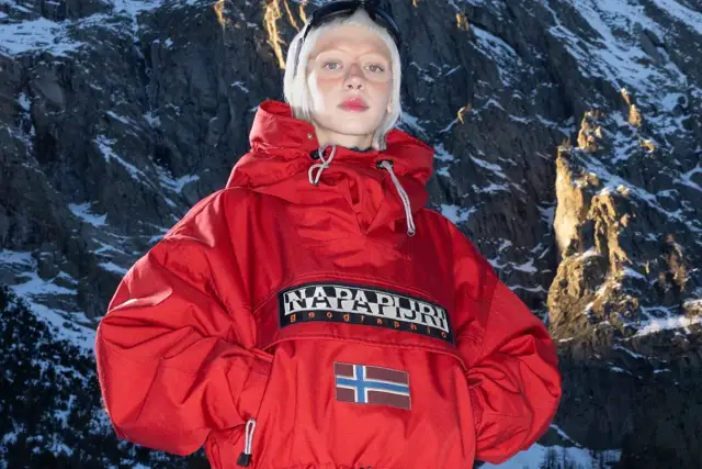 Modelka w czerwonej kurtce z logo Napapijri Geographic i flagą Norwegii na tle ośnieżonych gór. Napapijri co to za firma? To marka odzieży outdoorowej.