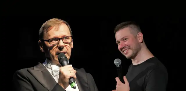 Dwóch mężczyzn z mikrofonami na scenie. Jeden w garniturze, drugi w czarnej koszulce. Widać, że socha stand up.