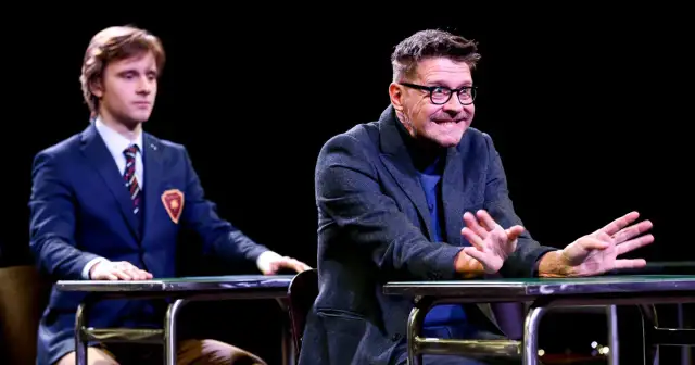 Dwóch aktorów na scenie podczas spektaklu "Stowarzyszenie Umarłych Poetów". Młody mężczyzna w garniturze i starszy z okularami, obaj siedzą przy stołach.