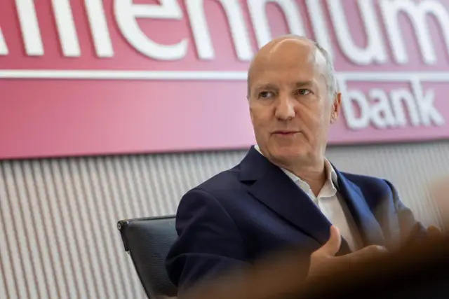 Mężczyzna w garniturze siedzi przed logo Banku Millennium. Kto jest właścicielem banku Millennium?