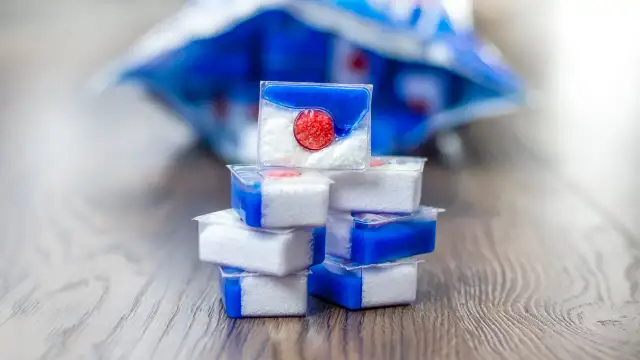 Stos kapsułek do zmywarki z niebieskim żelem i białą zawartością na drewnianym tle.