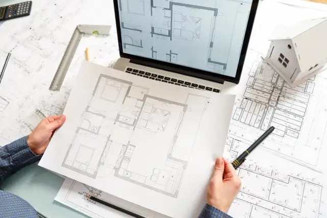 Ręce trzymające plan mieszkania, obok laptop z projektem, makieta domu i dokumentacja techniczna.