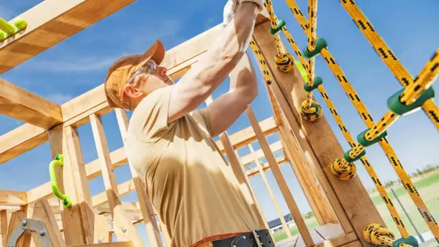 Budowa drewnianego placu zabaw: mężczyzna montuje żółte liny z zielonymi uchwytami.