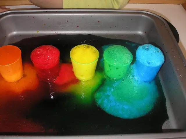 Kolorowe kubeczki z pianą w tacy, tworzące tęczę z płynów.