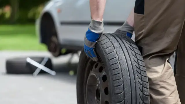 Mężczyzna w rękawiczkach trzyma koło zapasowe, przygotowując się do jego zamocowania w bagażniku. Obok leży klucz.