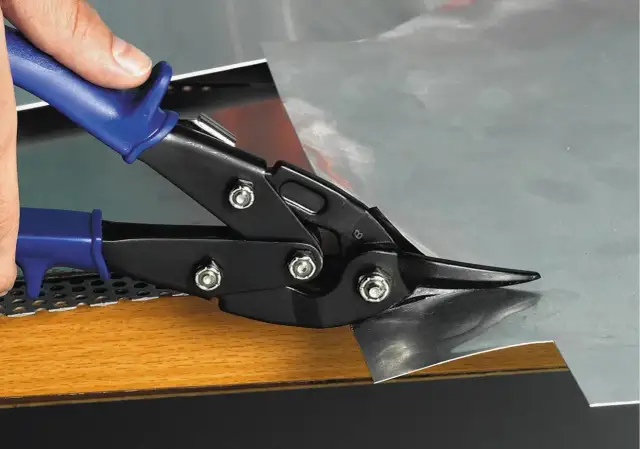 Dłoń trzyma niebieską rękojeść nożyc do blachy, które tną arkusz metalu.