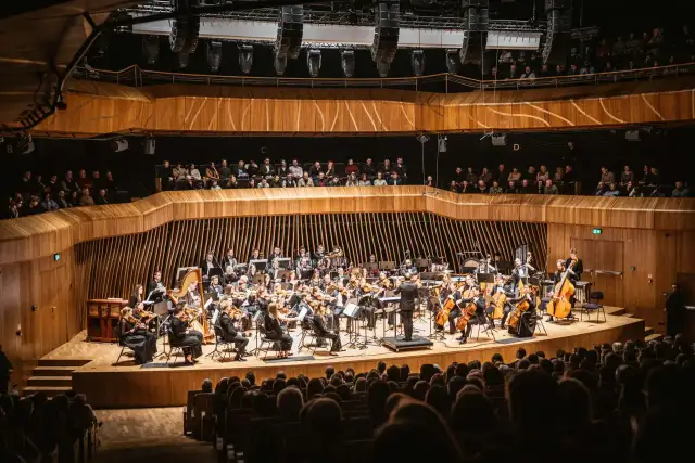 Orkiestra symfoniczna grająca w nowoczesnej sali koncertowej, z widownią na pierwszym planie.