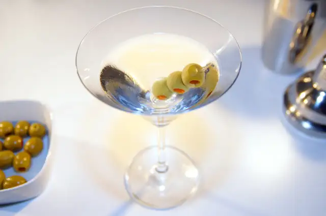 Kto wybierze Dirty Martini czy klasyczne Dry Martini? Kieliszek z oliwkami i drinkiem na białym tle.