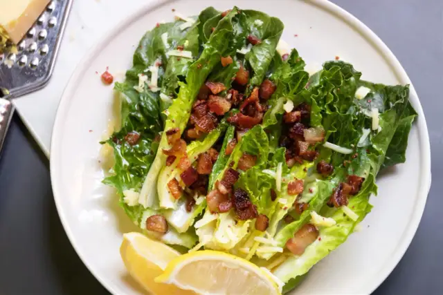 Sałatka cezar z boczkiem, chrupiące liście sałaty rzymskiej, posypane startym parmezanem i kawałkami chrupiącego boczku. Podana z cytryną.
