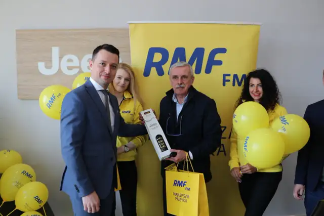 Uroczyste wręczenie nagrody w ramach finału loterii RMF FM. Mężczyzna w garniturze odbiera prezent od uśmiechniętej kobiety w żółtej kurtce.