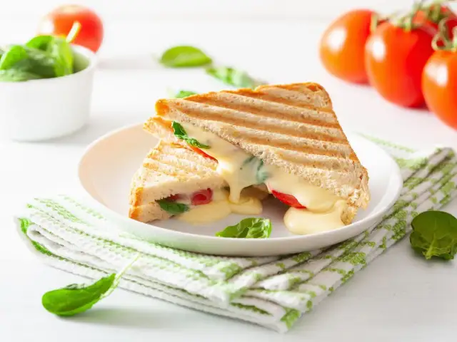 Grillowany tost z serem, pomidorami i szpinakiem na białym talerzu, obok pomidory i miseczka szpinaku.