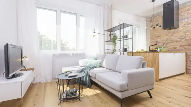 Jasny, przytulny pokój 15m2 z szarą kanapą, stolikiem kawowym, telewizorem i aneksem kuchennym z ceglaną ścianą.