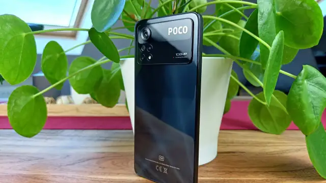 Czarny smartfon POCO X4 z potrójnym aparatem 108MP, otoczony zielonymi liśćmi rośliny.