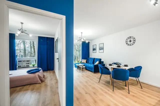 Nowoczesny pokój nad morzem z niebieskimi akcentami. Widok na salon z sofą i jadalnią oraz sypialnię z łóżkiem.