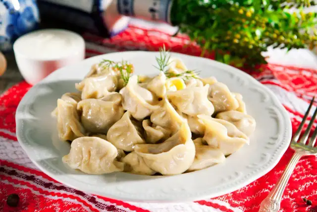 Pyszne uszka bezglutenowe przepis na talerzu, udekorowane koperkiem. Obok miseczka śmietany i widelec.