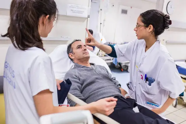 Lekarka bada pacjenta leżącego w łóżku szpitalnym, drugą pielęgniarkę widać z boku.