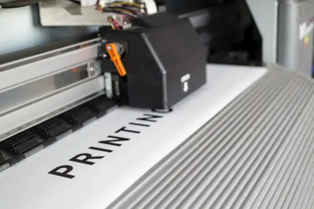 Głowica drukująca drukarki wielkoformatowej drukuje napis "PRINTING" na białym papierze.