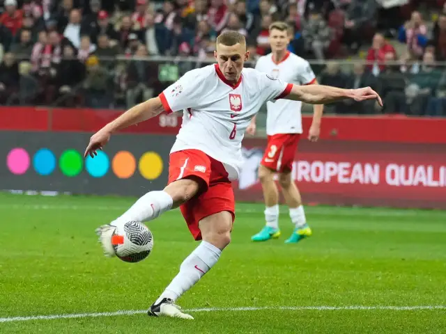 Piłkarz w białej koszulce z orłem i czerwonych spodenkach kopie piłkę na boisku.