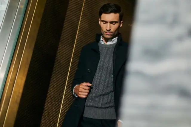 Mężczyzna w eleganckim swetrze jak marynarka, z warkoczowym splotem, w ciemnej, stylowej stylizacji.