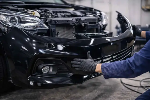 Mechanik w czarnej rękawiczce przykłada czarny zderzak do przodu samochodu.