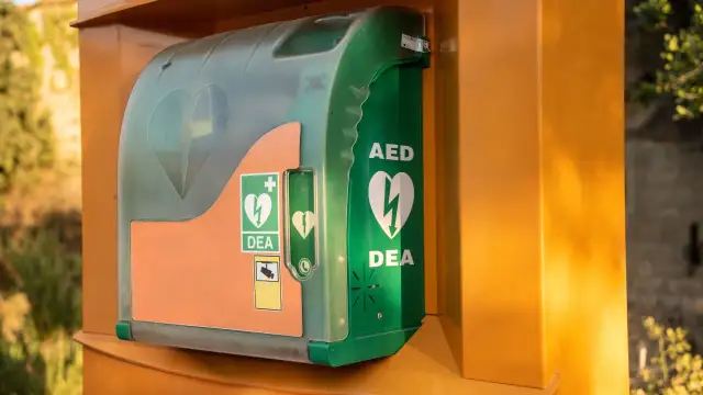 Zielona obudowa AED (defibrylatora) z symbolami serca i błyskawicy, zamontowana na pomarańczowym słupku.