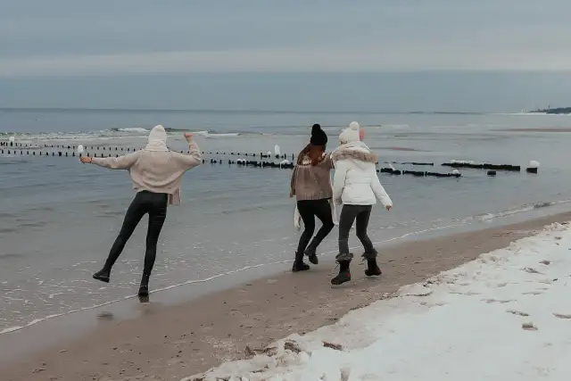 Trzy dziewczyny cieszą się zimą nad morzem, spacerując po zaśnieżonej plaży. Wesoła atmosfera i zimowy krajobraz.