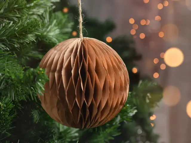 Brązowa, papierowa ozdoba choinkowa w kształcie kuli wisi na gałęzi choinki.