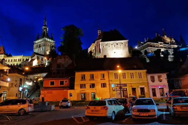 Nocne, oświetlone budynki i wieża zegarowa w Sighișoara. Atrakcje miasta nocą, z samochodami na pierwszym planie.