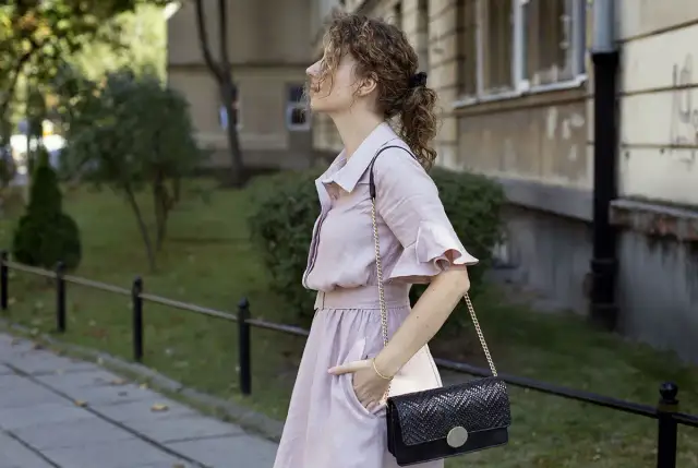 Kobieta w różowej sukience i czarnej torebce na łańcuszku na tle budynku i zieleni.