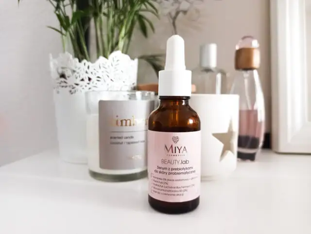 Brązowa buteleczka serum Miya Beauty.lab z pipetką na jasnoróżowej etykiecie, na tle świecy i roślin.