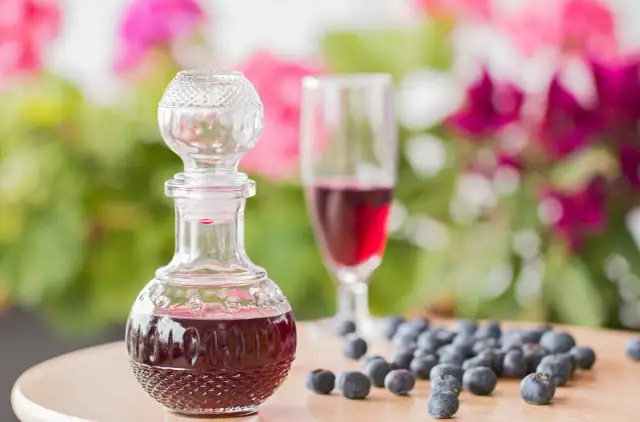 Elegancka karafka z nalewką z borówki, obok kieliszek i garść świeżych owoców. W tle kwitnące kwiaty.