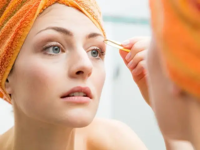 Kobieta z pomarańczowym ręcznikiem na głowie wyrywa brwi pęsetą, zastanawiając się, ile odrastają brwi.