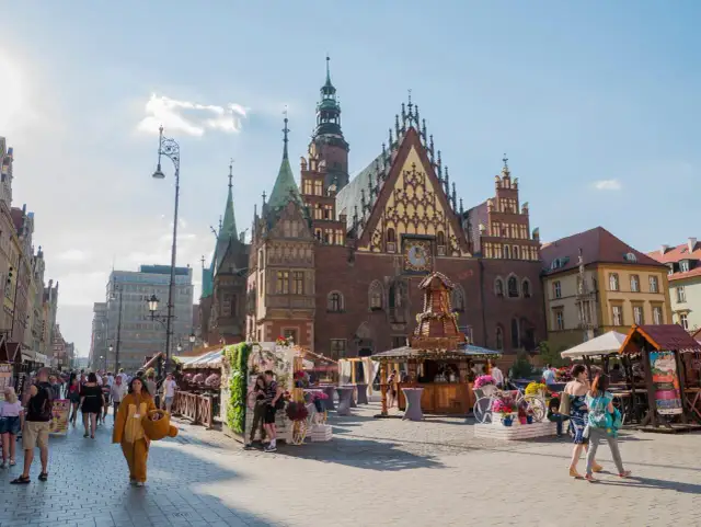 Tłum ludzi spaceruje po Rynku we Wrocławiu, gdzie można pojechać w niedzielę. W tle widać piękną architekturę i kramy z kwiatami.