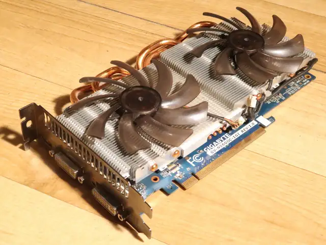 Karta graficzna Gigabyte GTX 560 Ti z dwoma wentylatorami i miedzianymi rurkami cieplnymi na drewnianym tle.
