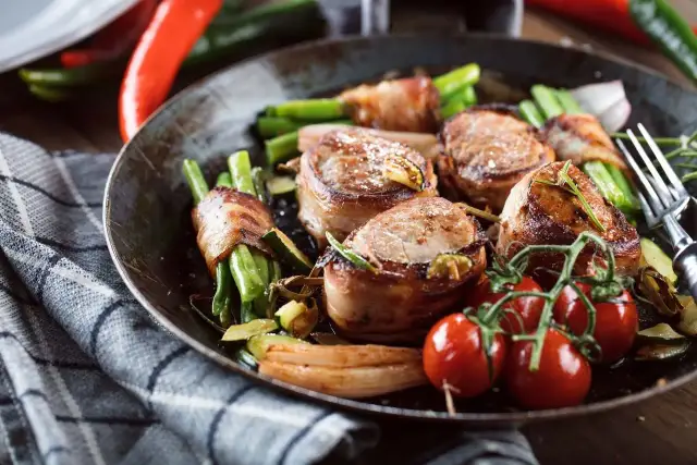 Polędwiczki wieprzowe zawinięte w boczek z fasolką szparagową i pomidorkami koktajlowymi na patelni.