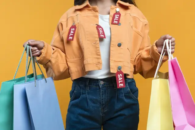 Kobieta w pomarańczowej kurtce z metkami "SALE" trzyma torby na zakupy. Symbol szybkiej mody i wyprzedaży.