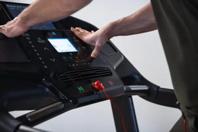 Dłonie użytkownika na panelu sterowania bieżni z widocznym klipsem bezpieczeństwa w kształcie serca.