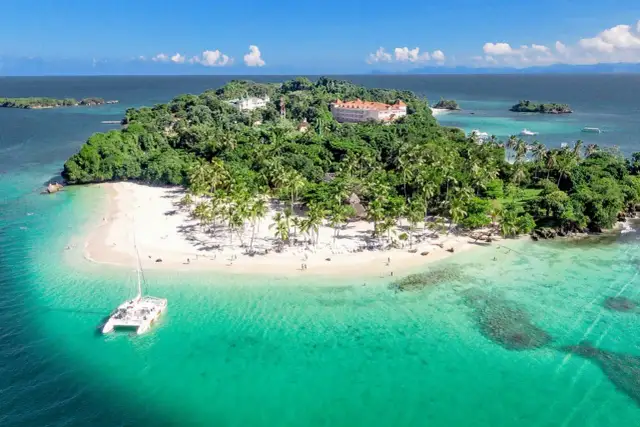 Rajska wyspa z białym piaskiem, palmami i hotelem. Idealne miejsce, by zobaczyć, co zwiedzić na Dominikanie. Katamaran na turkusowej wodzie.