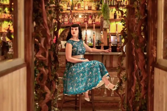 Kobieta w turkusowej sukience z koronki siedzi przy barze, opierając się o blat.