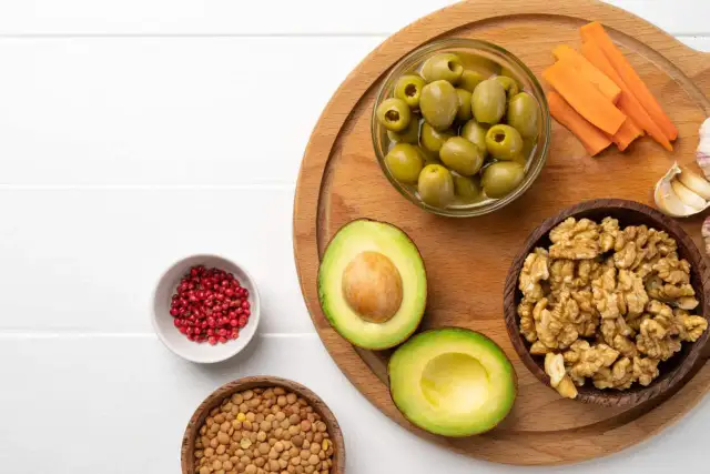 Awokado, orzechy, oliwki – to przykłady, jakie są tłuszcze roślinne. Na desce także marchewka, czosnek i różowy pieprz.