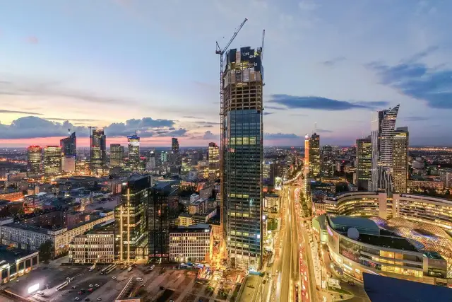 Warszawski wieżowiec w budowie na tle zachodzącego słońca i panoramy miasta.