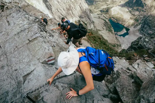 Grupa turystów wspina się po skalnej ścianie z pomocą łańcuchów, widok na jezioro w dole.