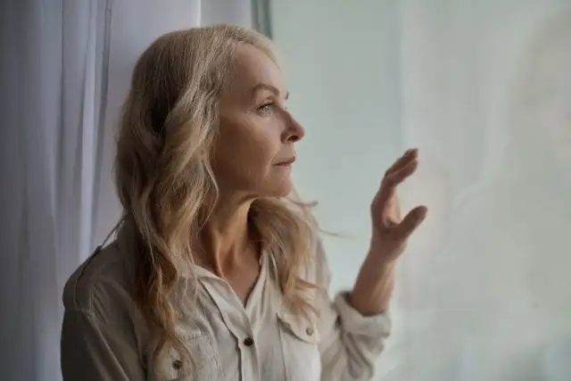 Starsza kobieta z blond włosami patrzy przez okno, zamyślona.